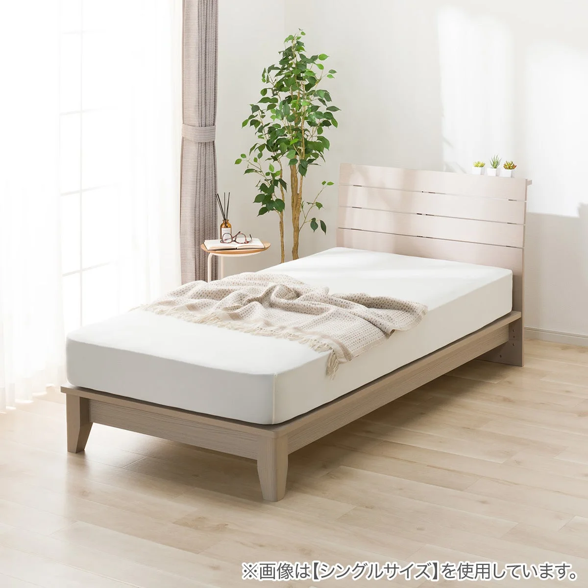 明るい日差しが差し込む部屋に、木目調のフレームが特徴的なシングルベッドが置かれています。白い寝具にベージュのブランケットがかけられ、ベッドサイドには観葉植物と小さなサイドテーブルがあり、リラックスできる空間を演出しています。